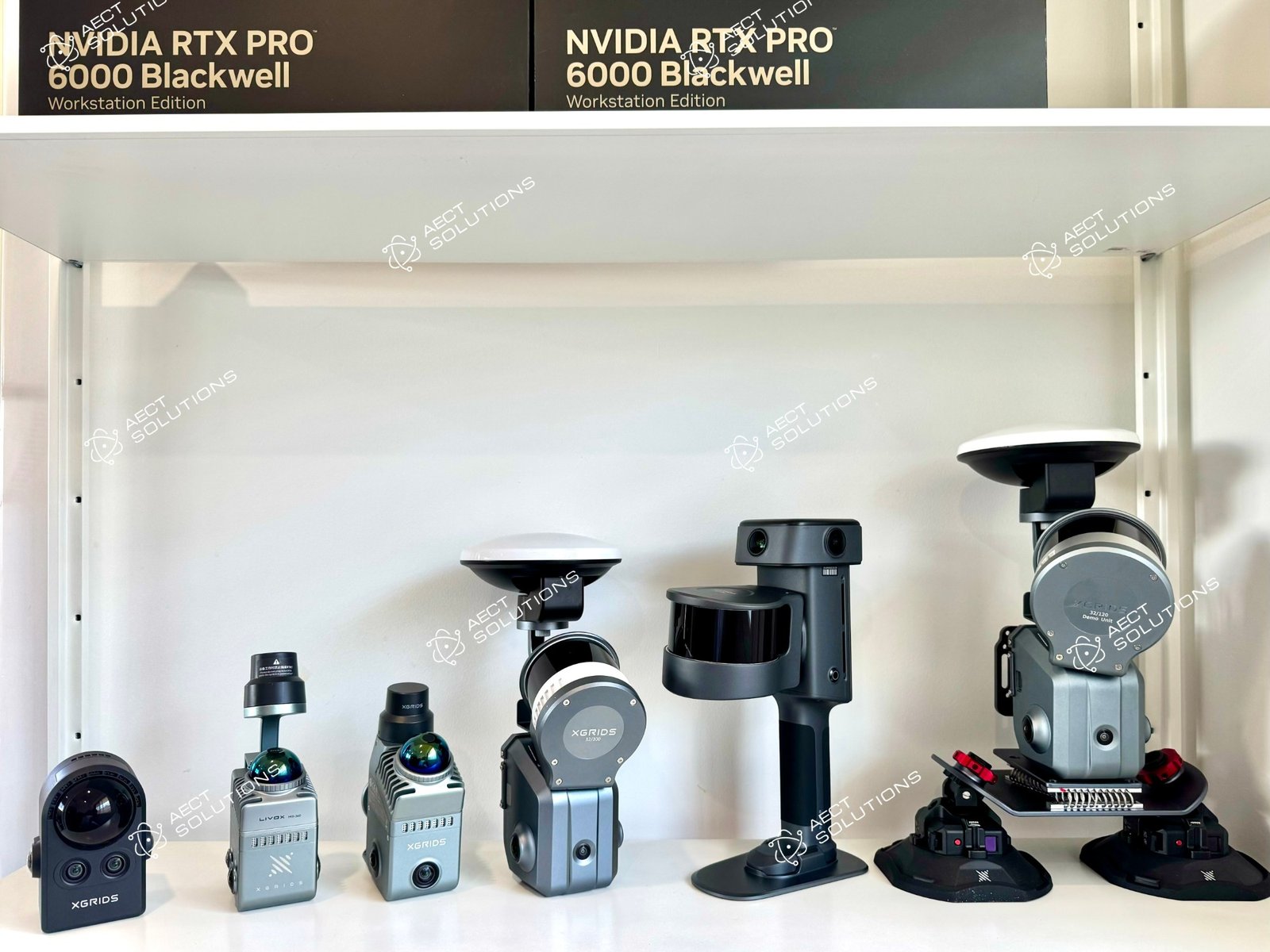 The full XGRIDS range on the AECT shelf in Queensland, left to right: PortalCam, Lixel K1, Lixel K2, Lixel L2 Pro 32-channel 300 m with RTK, the new Lixel L3, and an L2 Pro 32/120 on the vehicle mount, with NVIDIA RTX PRO 6000 workstation boxes above